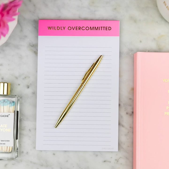 Notepad with 'Wildly Overcommitted' text, gold pen, and perfume bottle on a marble surface