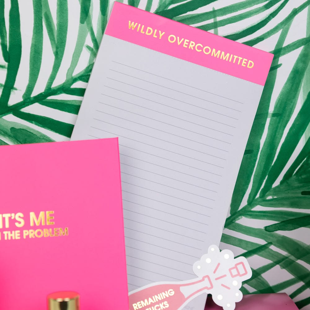 Notebook with 'Wildly Overcommitted' on a pink cover, surrounded by green leaves.