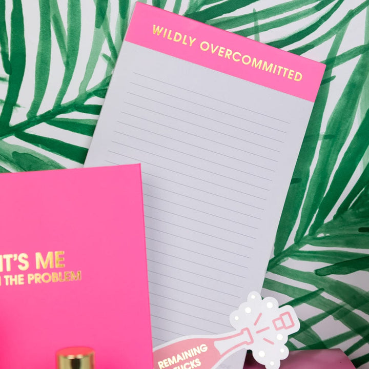 Notebook with 'Wildly Overcommitted' on a pink cover, surrounded by green leaves.
