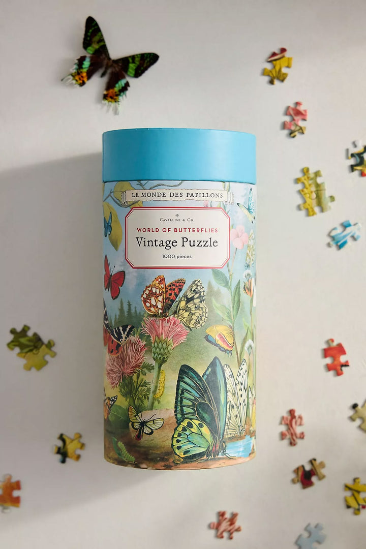 A cylindrical puzzle tin labeled 'Vintage Puzzle' with a light blue lid and a vintage-style butterfly and floral illustration, photographed on a white background with scattered puzzle pieces around it.